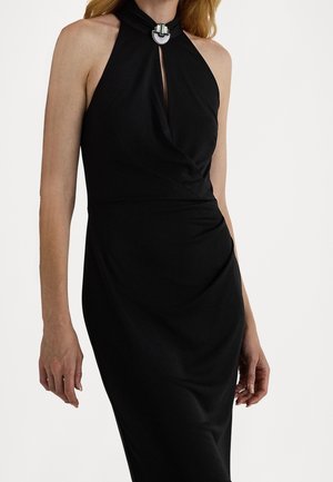 Žena v černých šatech bez rukávů s vykrojeným výstřihem a stříbrnou ozdobnou sponou u krku.