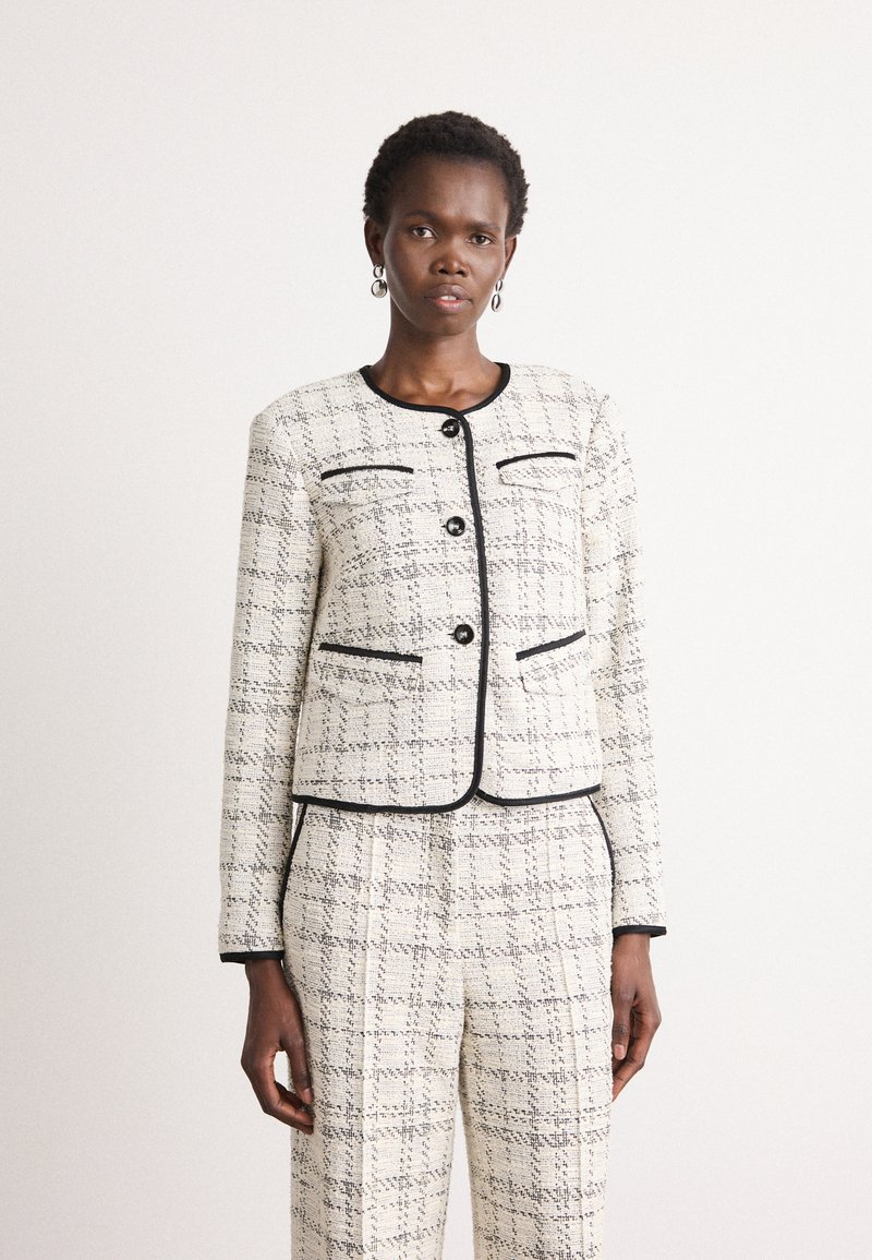 Veste en tweed blanche et noire avec un design court, un col rond et des poches avant. Présente des finitions noires et des boutons de fermeture.
