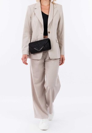 Femme portant un costume beige avec un pantalon large, un crop top noir, des baskets blanches et un sac bandoulière noir à bandoulière chaîne marchant vers l'avant.