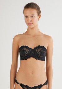 Brassiere negro sin tirantes con encaje que presenta patrones florales, bordes curvados, tela transparente y un diseño ajustado con soporte de aros.