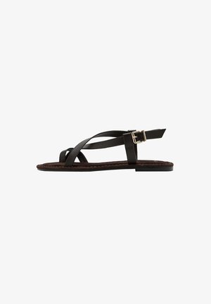 Sandale plate noire avec des lanières croisées et une bride à boucle dorée à la cheville, présentée de profil sur fond blanc.