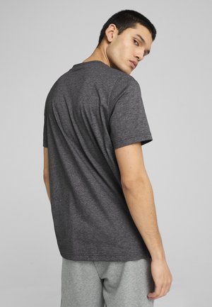 Młody mężczyzna w ciemnoszarej koszulce z krótkim rękawem i jasnoszarych spodniach, spoglądający przez ramię na jednolitym jasnym tle.