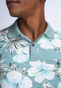 Lyhythihainen polo-paita vaaleansinisellä kukkakuviolla, jossa on kaulus, nappilista ja yksi rintatasku.