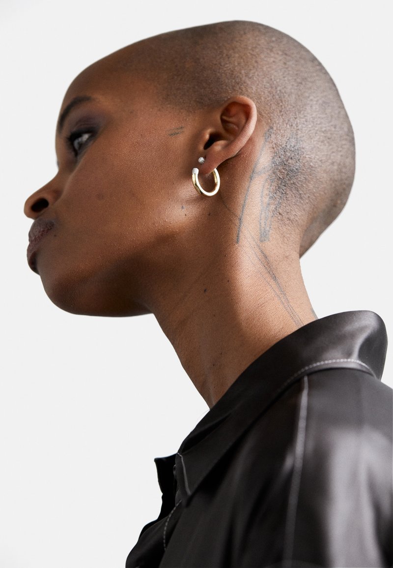 Black satin shirt with a collar, worn by a person with a shaved head. Features gold hoop and stud earrings, and a visible neck tattoo.