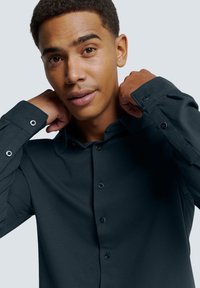 Donker, textuurgegeven overhemd met lange mouwen. Voorzien van ronde zwarte knopen en een klassieke kraag. Model poseert met handen op de kraag.