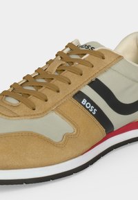 Καφέ και μπεζ δερμάτινο sneaker με καφέ κορδόνια, μαύρη ρίγα, λευκό λογότυπο "BOSS" και λευκή σόλα με κόκκινες λεπτομέρειες.