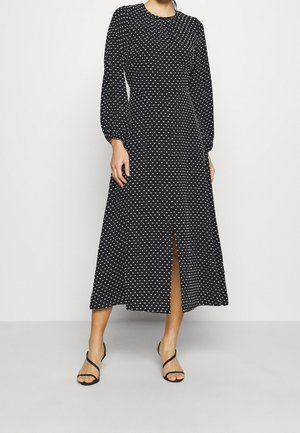 Robe midi noire à manches longues avec des pois blancs, une fente à l'avant et un col froncé, portée avec des sandales à talons noires à brides.