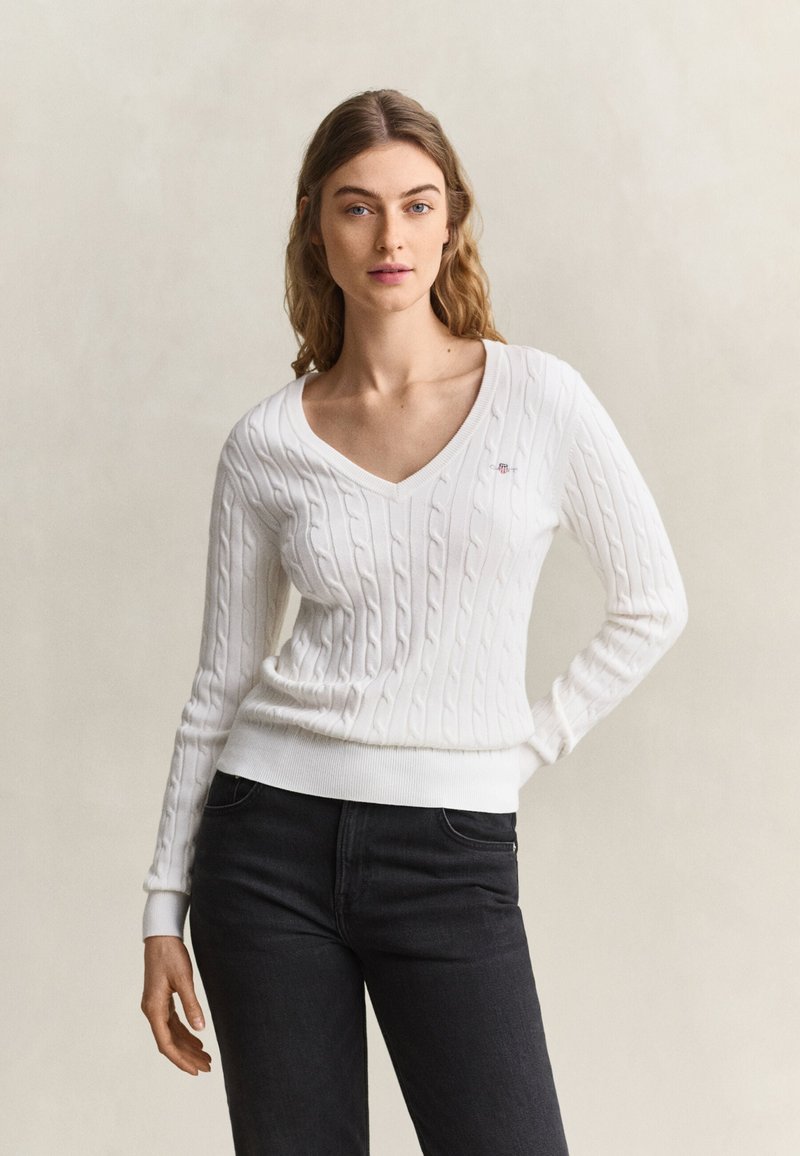 Jeune femme aux longs cheveux portant un pull blanc à col en V en maille torsadée et un jean noir, debout devant un fond clair uni.