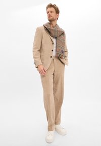 Beige Corduroy-Anzug mit einem taillierten Jacket und geraden Hosen, kombiniert mit einem gemusterten Schal in Rot, Schwarz und Weiß. Weiße Sneakers.