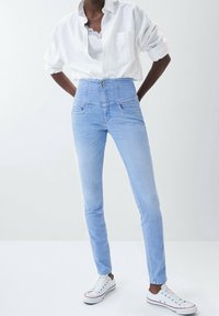 Jean bleu clair taille haute avec deux poches avant, fermeture à boutons, et coupe étroite aux jambes, assorti d'une chemise blanche boutonnée et de baskets.