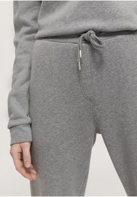 Grijze sweatpants met een trekkoord in de taille en knoopdetail, zachte textuur en taps toelopende benen, met een hand die ernaast rust.