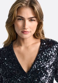 Crni top s sequinsima i V-izrezom, sa sjajnom, strukturiranom površinom i prilagođenim siluetom.