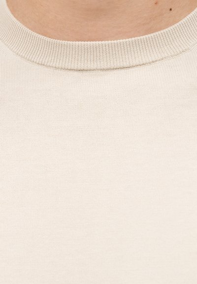 Κοντινή λήψη ενός ατόμου που φοράει μπεζ πλεκτό πουλόβερ με στρογγυλή λαιμόκοψη, εμφανίζοντας το λαιμό και το πάνω μέρος του στήθους.