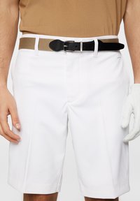 Białe szorty golfowe o gładkiej teksturze, zestawione z brązowym elastycznym paskiem i czarnym skórzanym akcentem. Szorty mają płaskie przednie kieszenie.