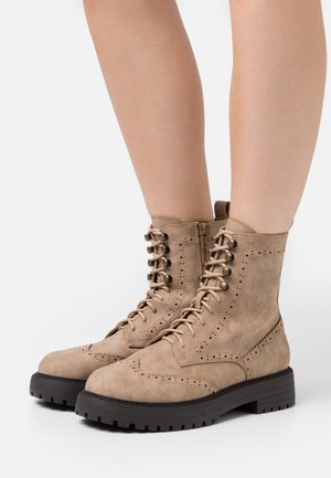 Botines de ante color camel con cierre delantero de cordones, ojales metálicos y detalles de brogue. Cuentan con una suela gruesa de goma negra y una cremallera lateral.