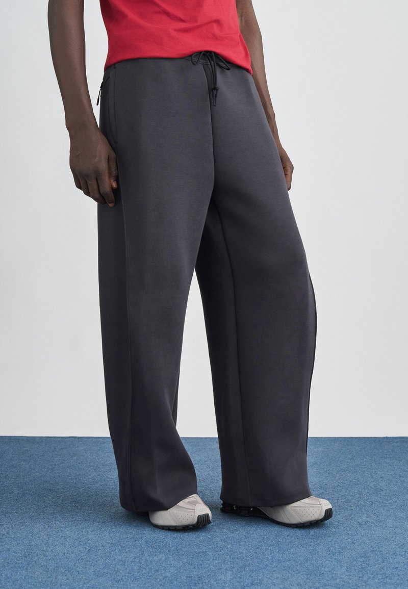 Pantaloni largi de culoare gri închis, realizați dintr-un material moale, cu talie reglabilă prin șnur și buzunare laterale cu fermoar. Asortați cu pantofi sport gri deschis.