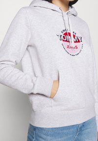Felpa grigia in misto cotone, dotata di tasca a marsupio frontale, cappuccio con coulisse e un audace design del logo "Tommy Jeans" sul petto.