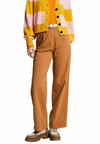 Femme portant un pantalon large marron, une ceinture blanche, des chaussures beiges épaisses, un haut orange et un cardigan rayé jaune-rose avec des boutons noirs.