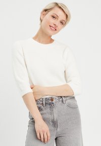 Pull en maille texturée blanc, à manches trois quarts, associé à un jean en denim gris clair taille haute avec fermeture à bouton.