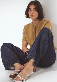 Ljussydd kortärmad tröja tillsammans med vida, mörkblå jeans. Vita sandaler med remmar och lågt klack. Enkelt, minimalistiskt design.