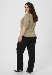 Mujer con cabello largo y castaño, vistiendo una blusa corta de mangas con brillo en color beige, pantalones negros y zapatos de tacón bajo negros, de pie y mirando hacia atrás.