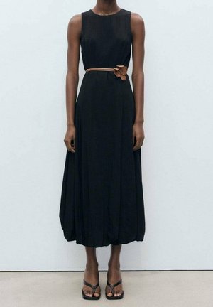 Kobieta w czarnej, bezrękawnej sukience midi z brązowym paskiem z kwiatową klamrą, w czarnych sandałach na otwartym palcu, stojąca na tle białej ściany.