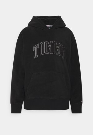 Person iført en sort hættetrøje med teksten "TOMMY", olivengrønne bukser med lap-detalje og hvide sneakers, står mod en ensfarvet hvid baggrund.