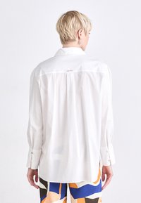 Camisa blanca de corte clásico con cuello, que presenta un pliegue central en la parte posterior y un sutil bordado decorativo. Mangas largas con puños de botón.