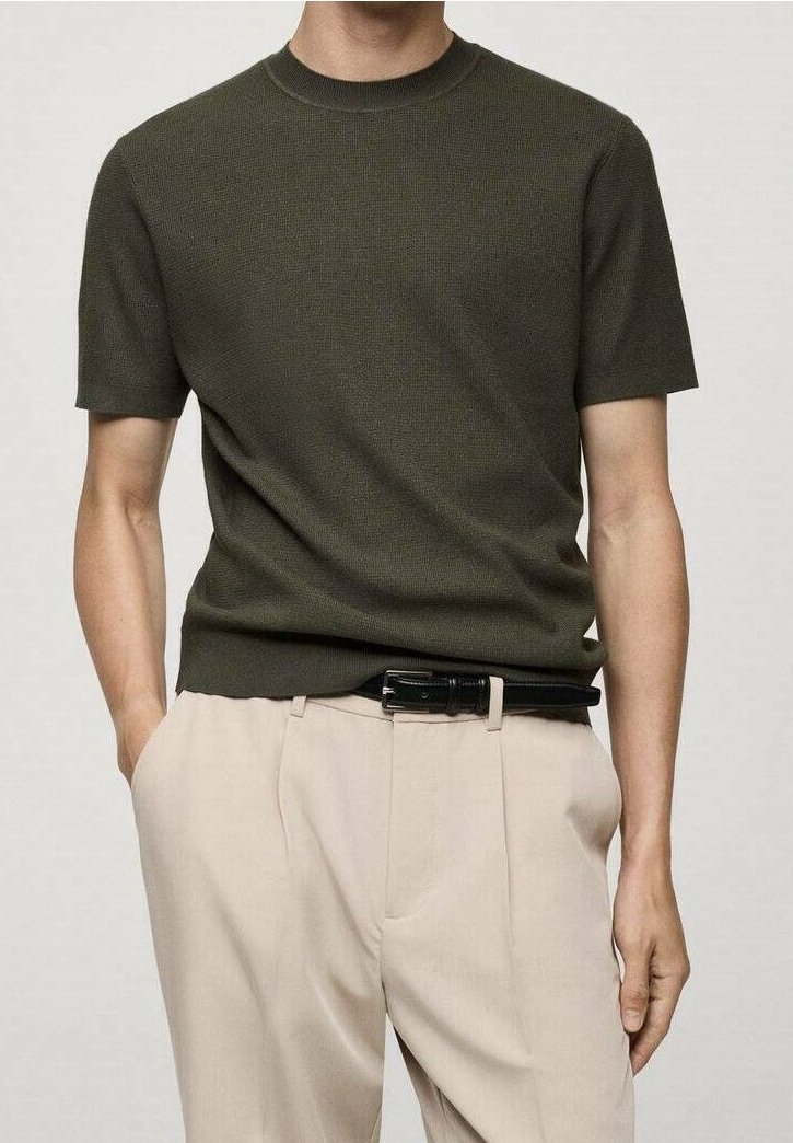 Homme portant un pull en maille à manches courtes couleur olive foncé, rentré dans un pantalon plissé beige avec une ceinture noire, mains dans les poches.