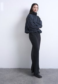 Νεαρή γυναίκα με μακριά σκούρα μαλλιά, που φοράει ένα μαύρο ριγέ φαρδύ σακάκι, μακριά μαύρη φούστα και μαύρες μπότες με τετράγωνη μύτη, μπροστά σε έναν λευκό τοίχο.