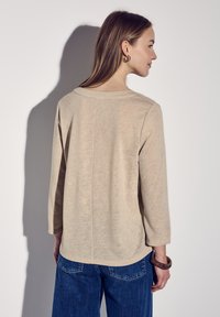 Beiger Pullover mit weitem Ausschnitt und dreiviertellangen Ärmeln, mit geradem Saum und subtiler Struktur. Kombiniert mit blauer Jeans.