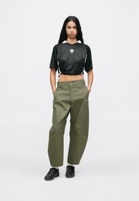 Mulher de pé com as mãos nos bolsos, vestindo uma blusa preta de manga curta e cropped, calças largas verdes-azeitona, meias brancas e sapatilhas pretas.