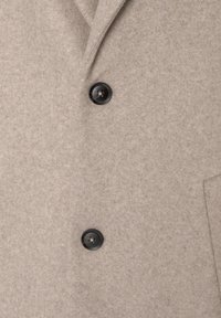 Section d'un manteau en laine beige montrant deux boutons noirs et une partie du col avec revers cranté.