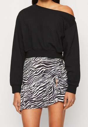 Kobieta ubrana w czarną, odsłaniającą ramiona, długorękawową bluzkę w połączeniu z krótką, zebra-printową spódnicą o marszczonym kroju, zakończoną falbaną z bocznym wiązaniem.