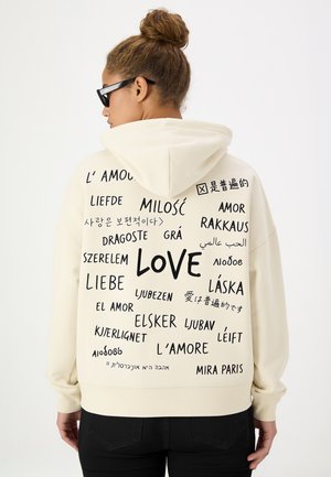 Personne portant un sweat à capuche blanc avec le mot "love" écrit en plusieurs langues dans le dos, debout devant un fond blanc uni.
