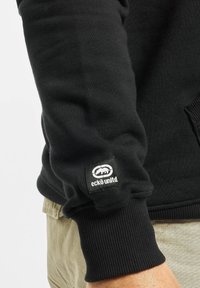 Sudadera negra con puños acanalados, que presenta una etiqueta cuadrada con un logo blanco y un diseño de oso en la manga izquierda.