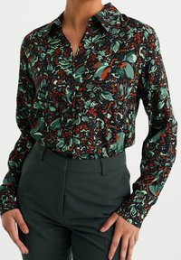 Kvinna i blus med långa ärmar och knappar med grönt och orange blommönster, instoppad i mörkgröna hög midja byxor.