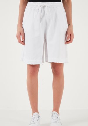 Žena oblečená v bielych šortkách dĺžky po kolená s pásom na šnúrku a nezačišteným lemom, doplnená bielymi teniskami a čiernym tričkom.