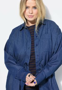 Camicia di jeans con vestibilità ampia, colore blu scuro, tasca frontale e cravatta regolabile in vita. Sotto c'è una maglietta a righe orizzontali.