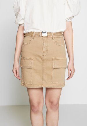 Beige denim mininederdel med forlommelommer og bælte, båret med en løs hvid top, vist fra taljen til knæene.