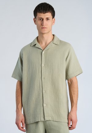 Młody mężczyzna z krótkimi ciemnymi włosami, ubrany w luźną, jasnozieloną, krótkomanicową koszulę na guziki oraz pasujące spodnie, stojący na jednolitym tle.