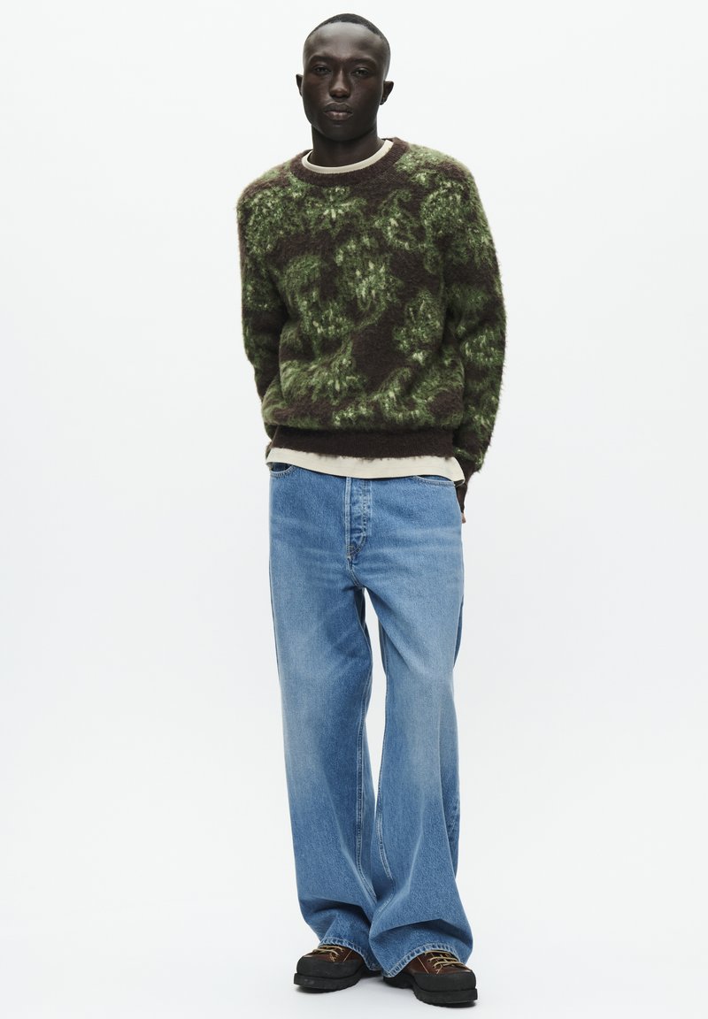 Strikkegenser i mørkeblå og grønn med blomstermønster, kombinert med lyseblå vide jeans og mørke sko. Uformell antrekkspresentasjon.