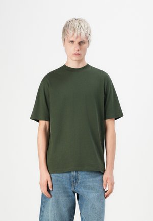Camiseta de algodón verde oscuro con cuello redondo y mangas cortas, combinada con unos jeans de mezclilla azul claro. La tela parece suave y lisa.