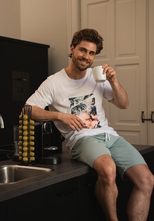 Witte T-shirt met een kleurrijke tropische grafiek, lichtblauwe shorts, zittend op een aanrecht naast een koffiemachine en capsulehouder.