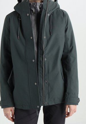 Persona con indosso una giacca con cappuccio verde scuro sopra una felpa grigia con zip, con pantaloni neri, in piedi su uno sfondo chiaro e uniforme.