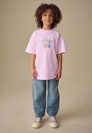 Kind mit lockigem Haar, das ein pastellrosa Hello Kitty T-Shirt, locker sitzende blaue Jeans und gemusterte Sneakers trägt und vor einem beigen Hintergrund steht.