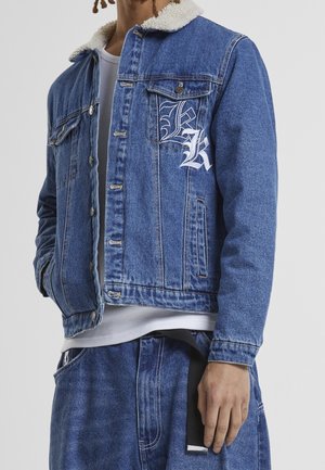 Personne portant une veste en jean bleue avec un col en polaire blanche et des lettres brodées "K" et "L" sur la poche poitrine, assortie à un jean assorti.