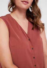 Top sans manches en rose poussiéreux avec décolleté en V, orné de boutons noirs et d'un léger éclat. Comprend une poche latérale et un tissu lisse et léger.