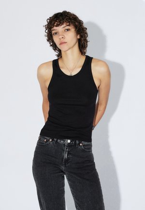 Personne aux cheveux courts et bouclés portant un haut sans manches noir et un jean foncé, debout devant un fond clair uni.