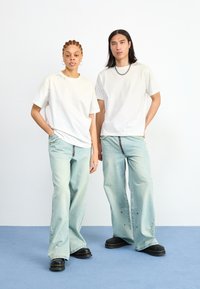 Dos modelos están uno al lado del otro, vistiendo camisetas blancas y jeans de pierna ancha azul claro con dobladillo sin terminar, combinados con zapatos de plataforma negros.